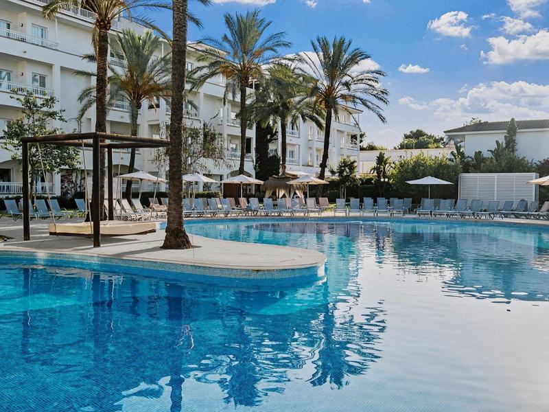 Großer blauer Pool mit Palmen, weißen Liegestühlen und modernem Hotelgebäude im Hintergrund.