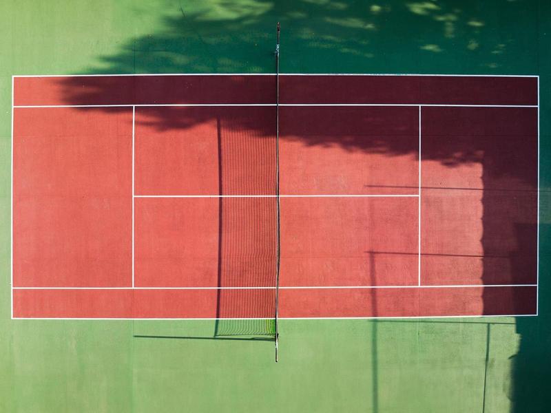 Tennisplatz aus der Vogelperspektive mit rotem Spielfeld und grüner Außenfläche.