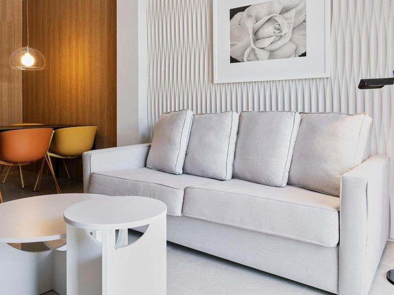 Hellgraues Sofa mit Kissen vor weißer Wand, rundem weißen Couchtisch, schwarzer Stehlampe und Stühlen.