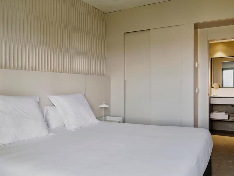 Schlichtes Schlafzimmer mit Bett, weißen Kissen, minimalistischer Wandgestaltung und Blick ins Badezimmer.