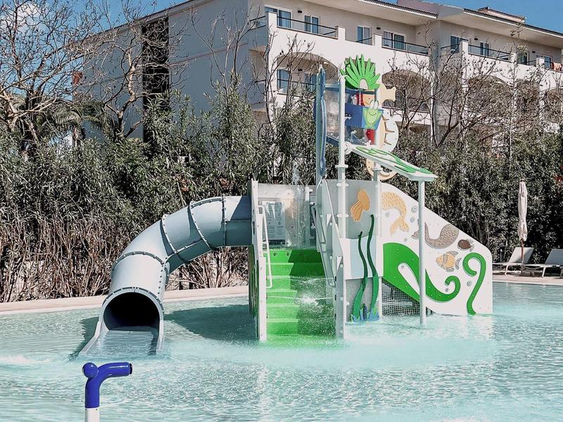 Kinderspielplatz mit grünem Wasserrutschen-Turm und blauem Röhrenrutsche in flachem Wasserbecken.