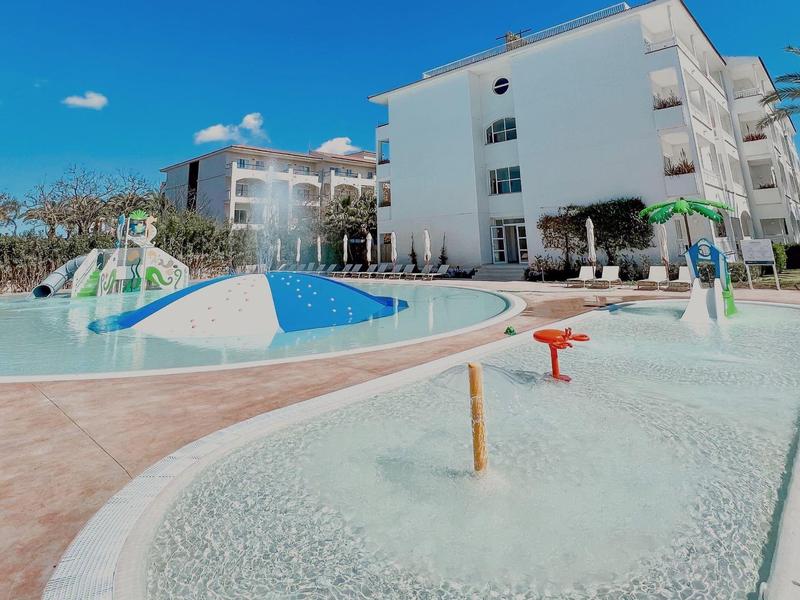 Wasserpark mit Planschbecken, Rutschbahn, Spritzspielzeugen und Wohnungen im Hintergrund bei Sonnenschein.