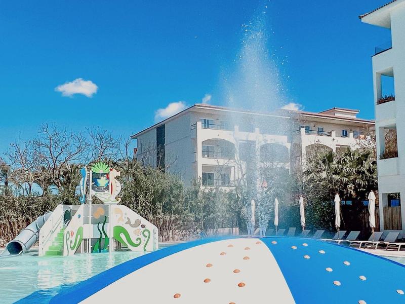 Hotelgebäude mit springbrunnen, grüner Rutsche und Wasserspiel im sonnigen Außenbereich.