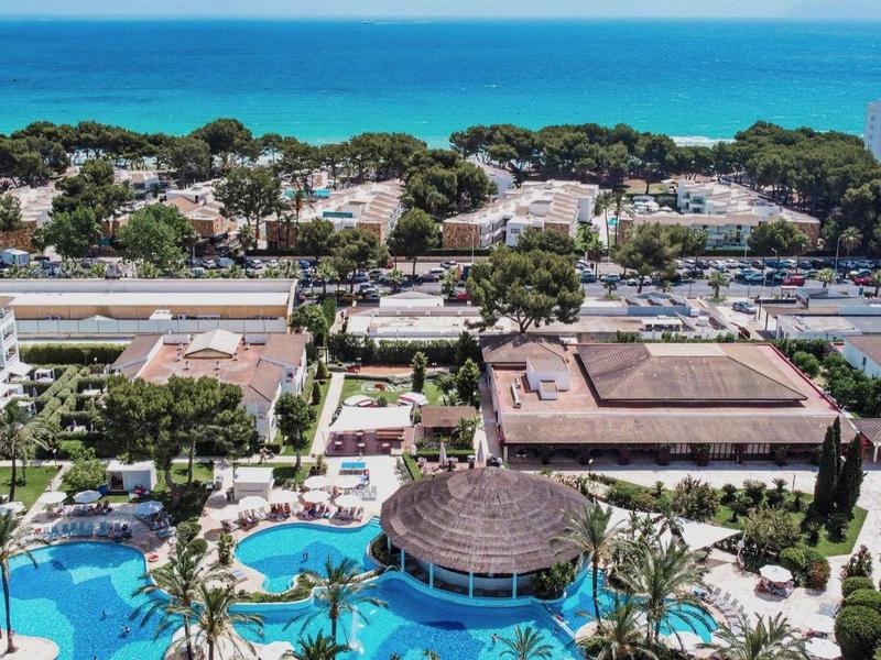 Hotelanlage mit Pool, Palmen und Blick aufs Meer, umgeben von Gebäuden und grünem Uferbereich.