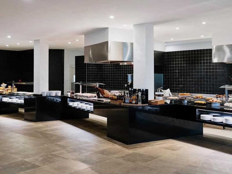 Moderne Großküche mit schwarzen Arbeitsflächen, Edelstahlgeräten und weißen Säulen unter Deckenbeleuchtung.