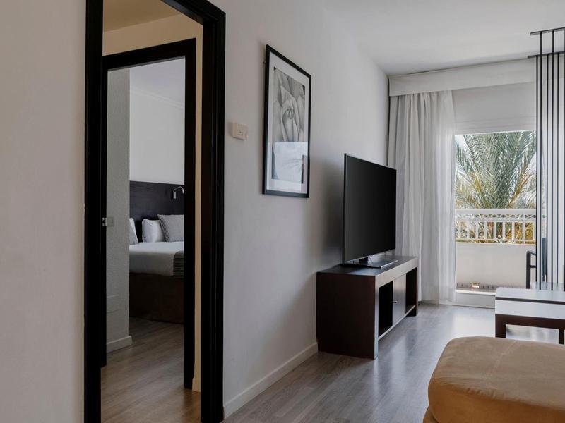 Moderner Wohnraum mit hellem Boden, schwarzer TV-Konsole, großem Fenster und angrenzendem Schlafzimmer.