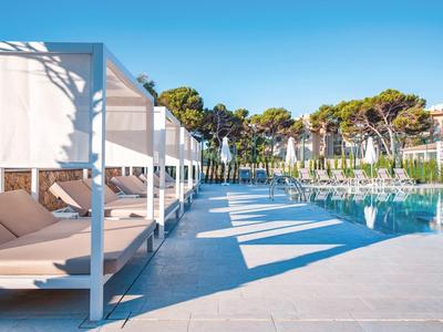 Area piscina moderna con lettini bianchi e alberi sullo sfondo sotto un cielo limpido.