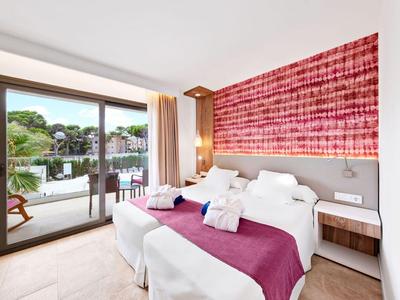 Chambre d'hôtel lumineuse avec deux lits, couvertures violettes, mur rouge et balcon avec vue sur le lac.