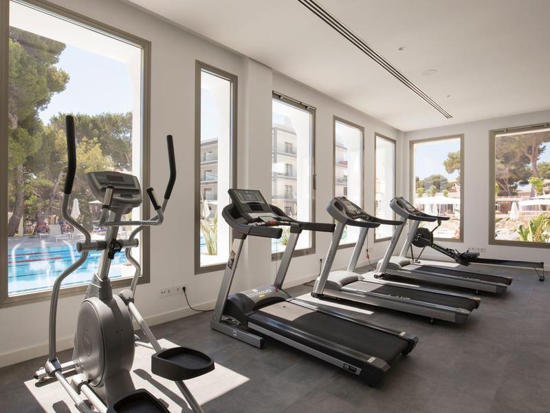 Salle de sport lumineuse avec tapis de course et elliptique près de grandes fenêtres donnant sur la piscine.