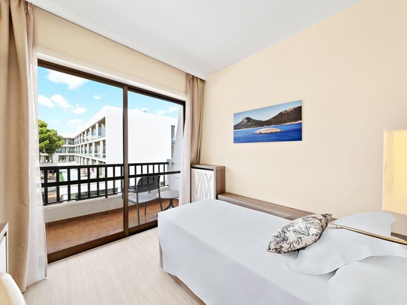 Chambre d'hôtel lumineuse avec lit simple, balcon et vue sur les bâtiments voisins.