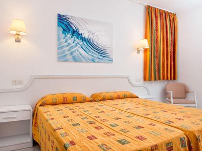 Pokój hotelowy z łóżkiem podwójnym, żółtą pościelą w wzory, stolikiem nocnym, fotelem i oknem z pomarańczową zasłoną.