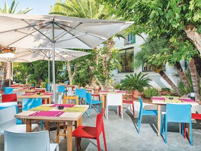 Bunte Stühle und Tische unter Sonnenschirmen in einem grünen, sonnigen Außenrestaurant.