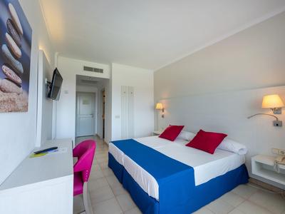 Nowoczesny pokój hotelowy z dwuosobowym łóżkiem, czerwonymi poduszkami i lampkami ściennymi w jasnym wnętrzu.