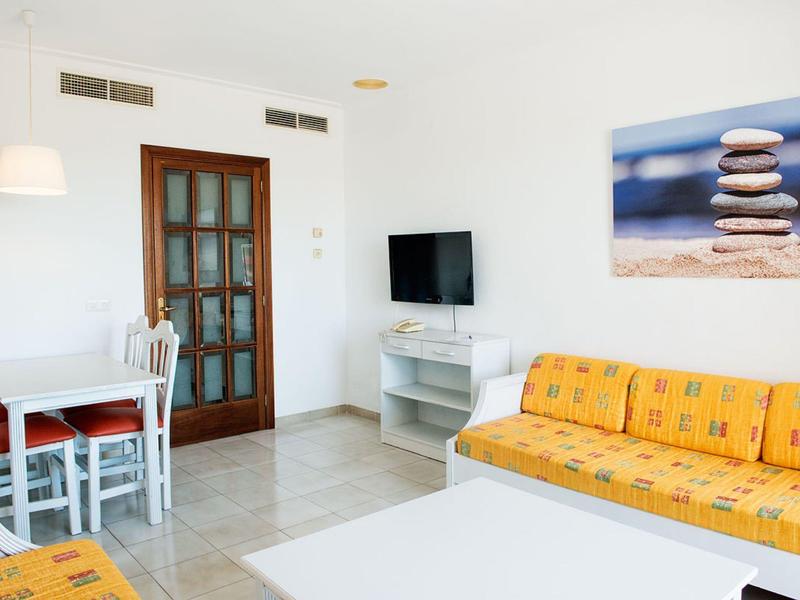 Jasny salon z żółtą sofą, małym stołem jadalnym, telewizorem na ścianie i obrazem kamieni.