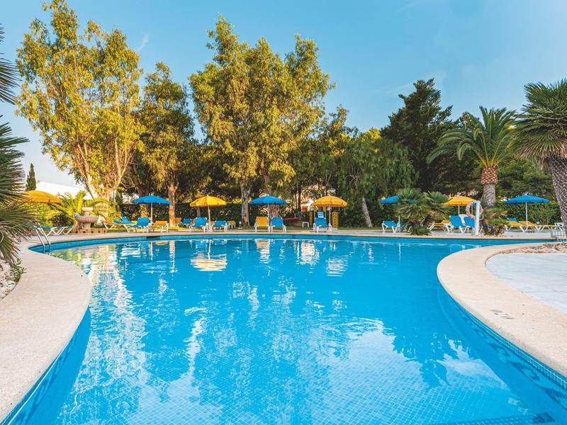 Basen z niebieskimi leżakami i parasolami otoczony drzewami i palmami.