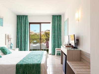 Camera d'hotel luminosa con letto matrimoniale, tende verdi e porta sul balcone con vista sul giardino.