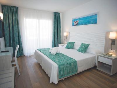 Camera d'hotel luminosa con letto matrimoniale, tende turchesi, copriletto, pavimento in legno e comodini.