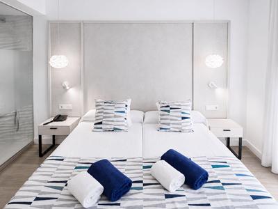 Nowoczesny pokój hotelowy z dwoma pojedynczymi łóżkami, białymi i niebieskimi poduszkami oraz szklaną prysznicem po lewej.