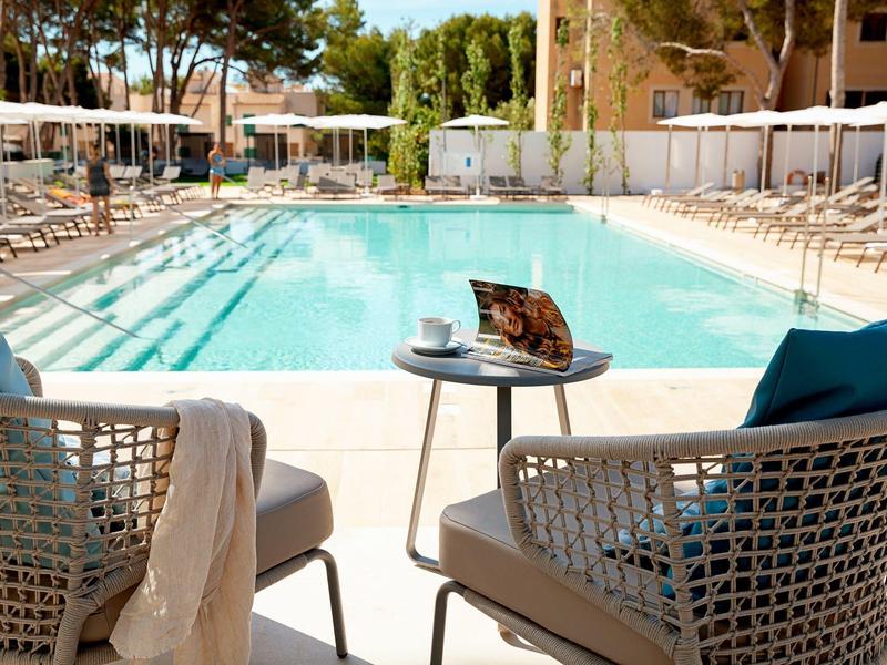 Piscina dell'hotel con sedie, tavolo, laptop e caffè circondata da lettini e ombrelloni.