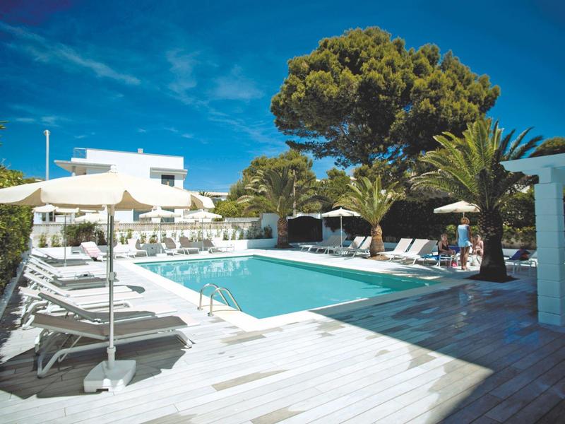 Area piscina moderna con lettini bianchi e ombrelloni sotto un cielo azzurro.