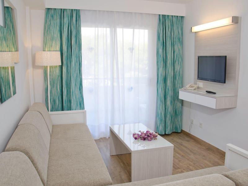 Soggiorno luminoso con divano, tavolino bianco, TV a schermo piatto e tende verdi.