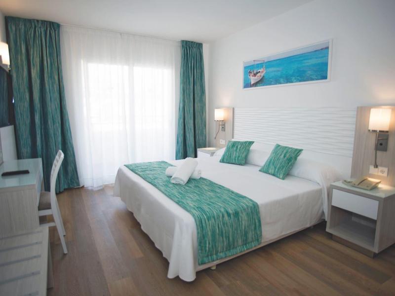 Camera d'hotel luminosa con letto matrimoniale, tende turchesi, copriletto, pavimento in legno e comodini.