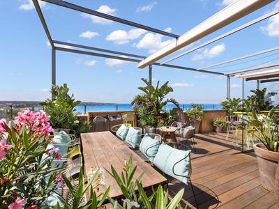 Terrazza sul tetto con tavolo in legno, cuscini, piante e vista sul mare sotto cielo sereno.