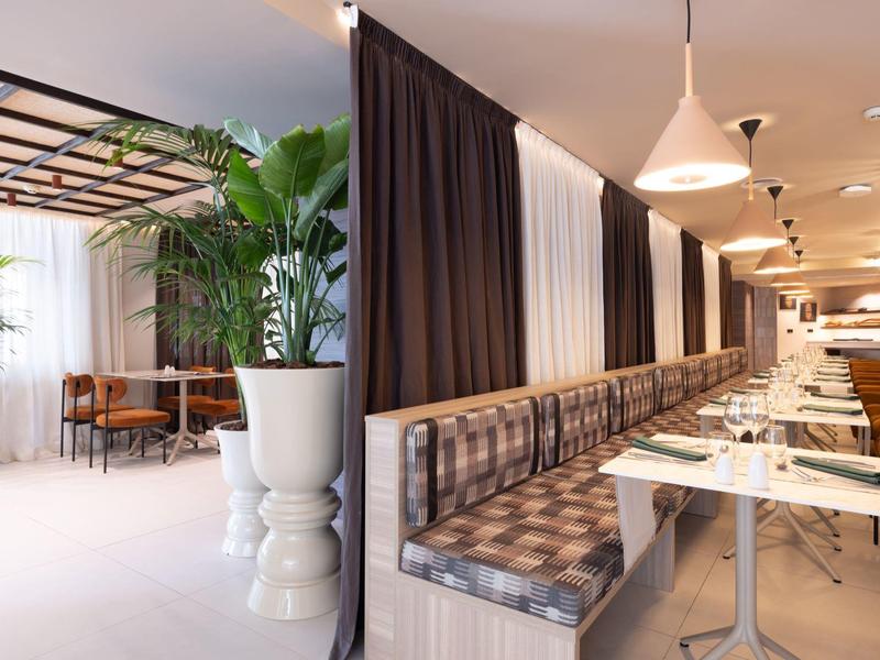 Lounge d'hotel moderna con piante alte, tende marroni e sedute a panchina con motivo a quadri.