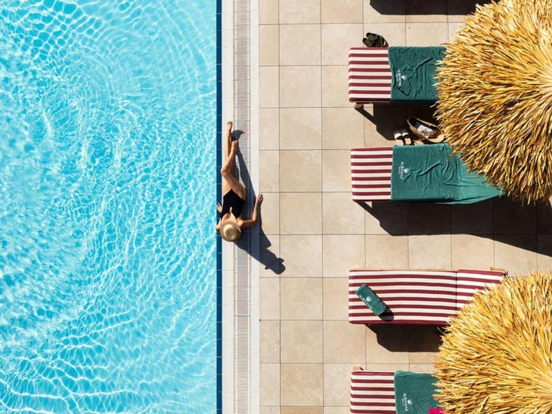 Vista aerea di un'area piscina con lettini e ombrelloni accanto ad acqua limpida.