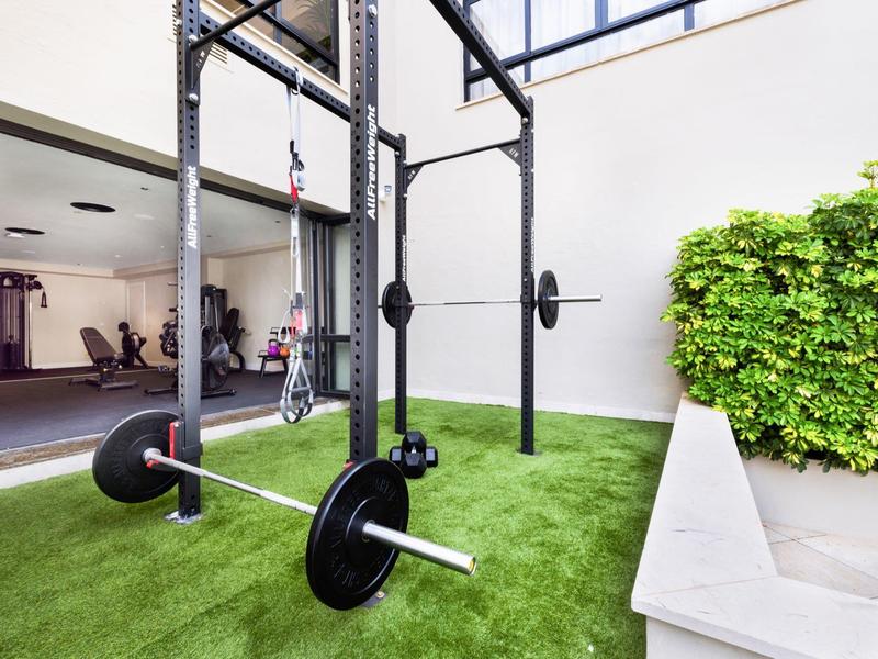 Area allenamento all'aperto moderna con attrezzature fitness su prato sintetico verde accanto a un muro bianco con piante.