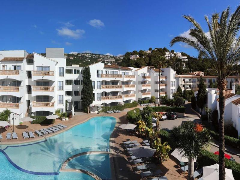 Complesso alberghiero con piscina, palme e edifici bianchi su più piani sotto un cielo soleggiato.