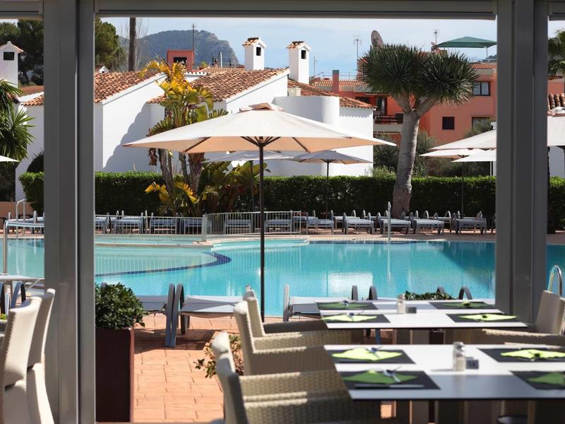 Vista da un ristorante su una piscina con ombrelloni bianchi e case sullo sfondo.