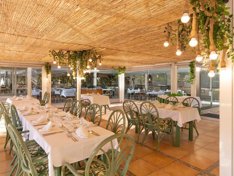 Hell beleuchteter Gastraum mit gedeckten Tischen, grünem Mobiliar und Pflanzen an der Decke.