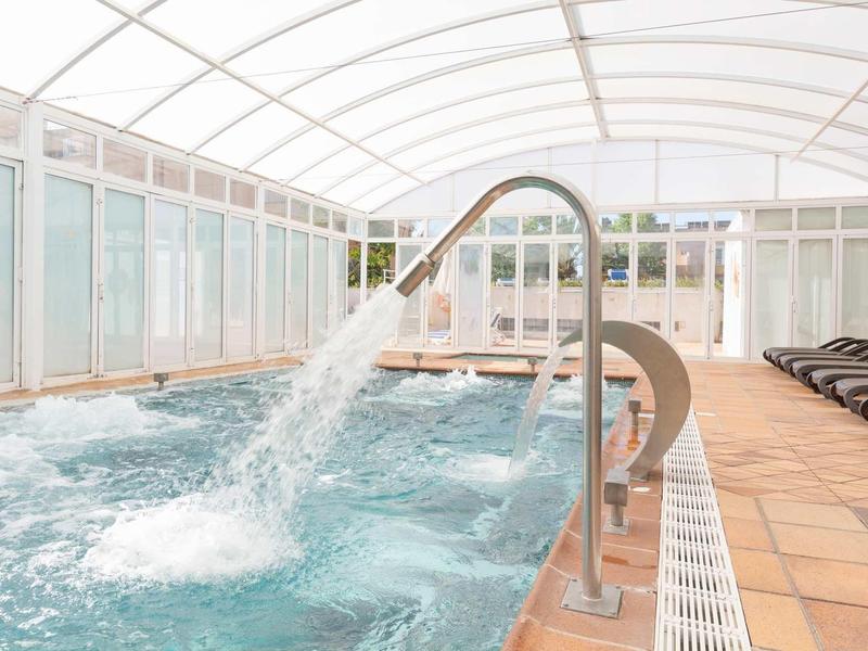 Innen-Schwimmbad mit großen Fenstern, Sprudelbecken und braunen Liegestühlen am Beckenrand.