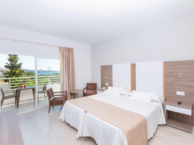 Helles Hotelzimmer mit Doppelbett, Holzmöbeln, Balkon und Blick auf grüne Landschaft.