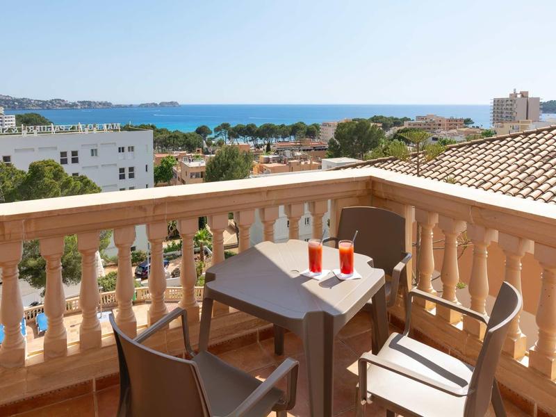 Balkon mit kleinem Tisch und zwei Stühlen, Sicht auf Meer und Gebäude bei klarem Himmel.