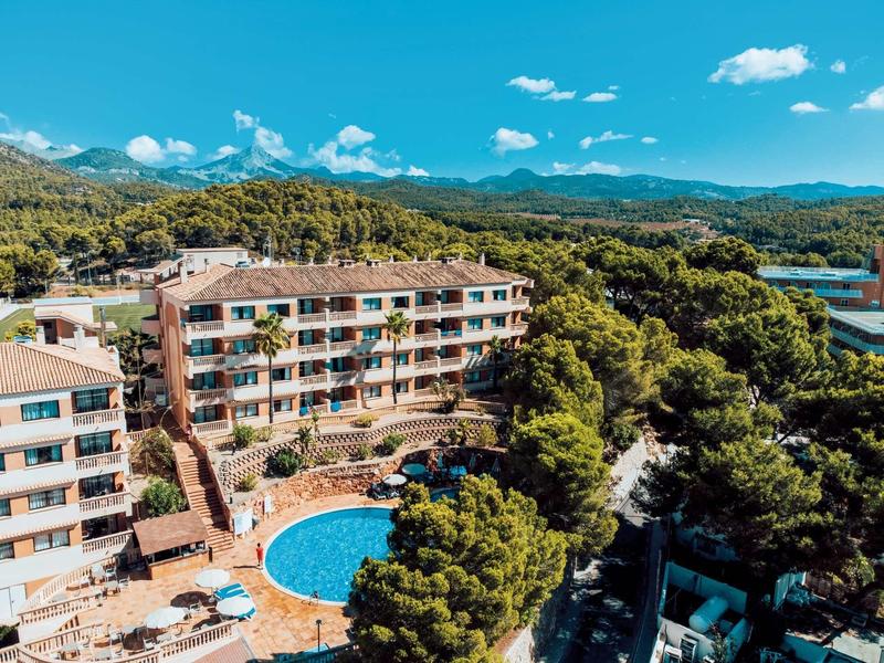 Mehrstöckige Wohnhäuser mit Rundpool, umgeben von grünen Bäumen und Bergen unter blauem Himmel.