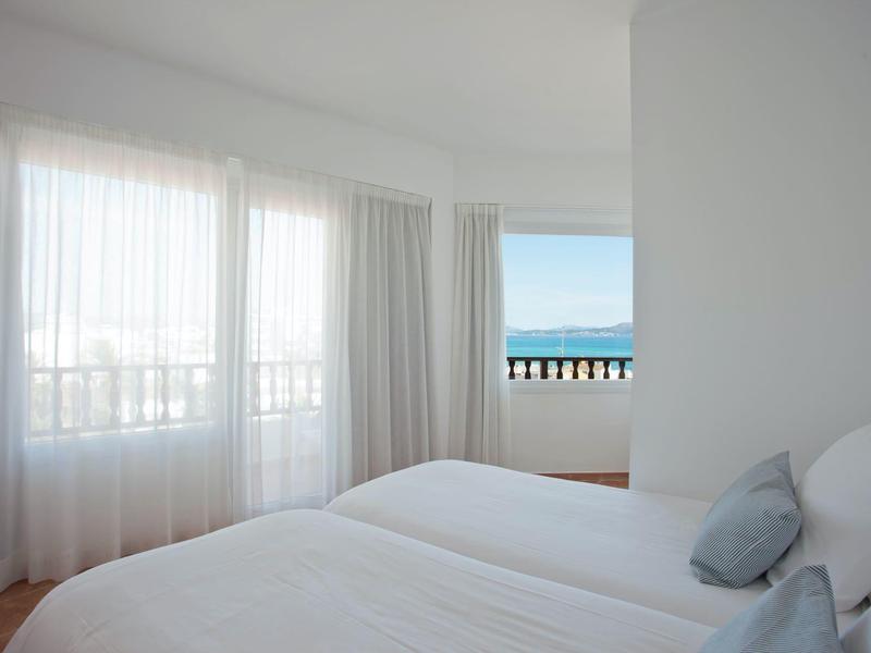 Jasna sypialnia z podwójnym łóżkiem, białymi zasłonami i widokiem na morze przez okno.