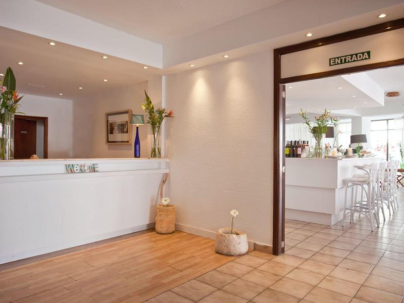 Jasna recepcja hotelowa z białym biurkiem, drewnianą podłogą i widokiem na przyległy kącik wypoczynkowy.