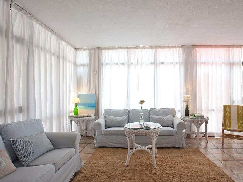 Jasny salon z szarymi sofami, okrągłym stołem i dużym oknem z białymi zasłonami.