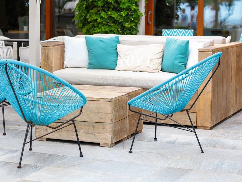 Área de asientos exterior moderna con mesa de madera, sofá y sillas turquesas sobre suelo de piedra.