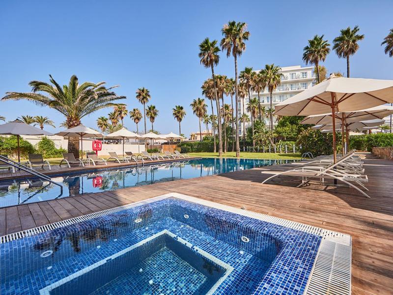 Área de piscina con tumbonas, sombrillas y palmeras en un resort hotelero bajo cielo despejado.