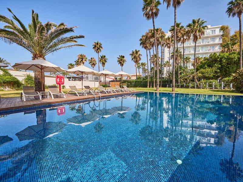 Piscina de hotel con azulejos azules, palmeras y tumbonas bajo cielo soleado.