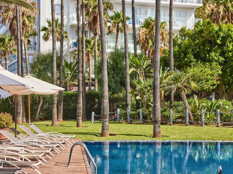 Piscina del hotel con tumbonas, sombrillas y palmeras cerca de un hotel.