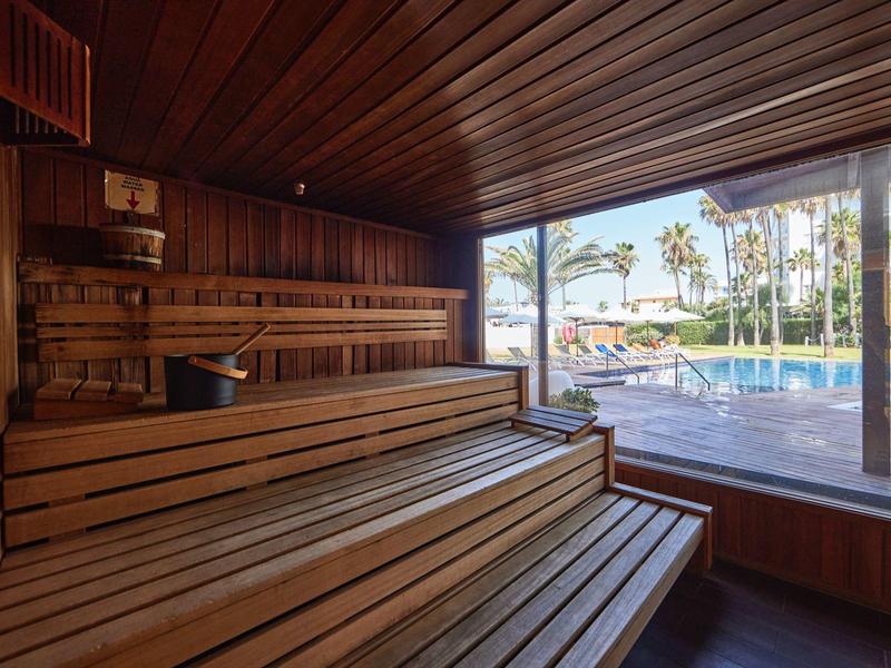 Sauna con bancos de madera y vista a través de grandes ventanas hacia una piscina y palmeras.