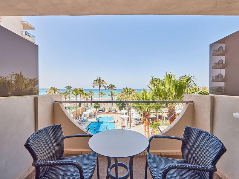 Balcón con dos sillas y mesa frente a una piscina y palmeras en un complejo hotelero junto al mar.