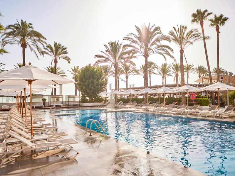 Área de piscina soleada con tumbonas y palmeras en un resort vacacional frente al mar.