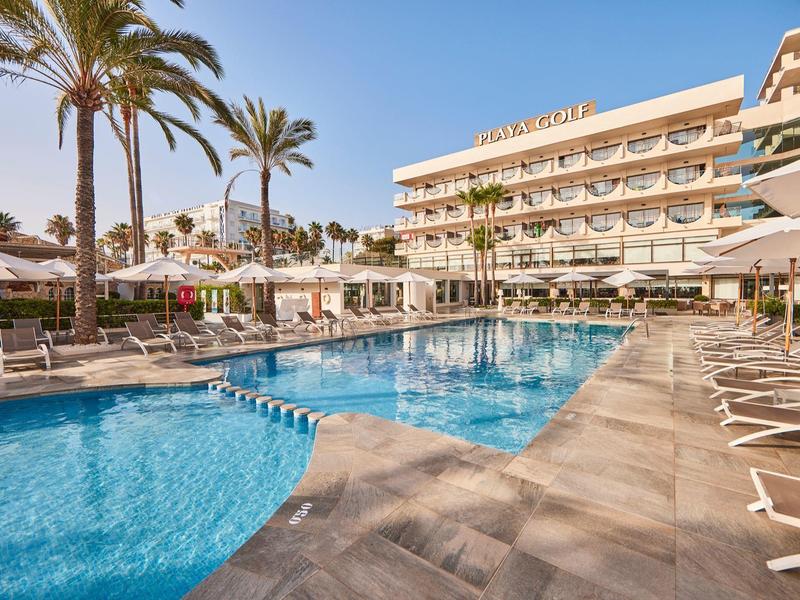 Hotel con gran piscina exterior, sombrillas y tumbonas bajo palmeras en cielo despejado.