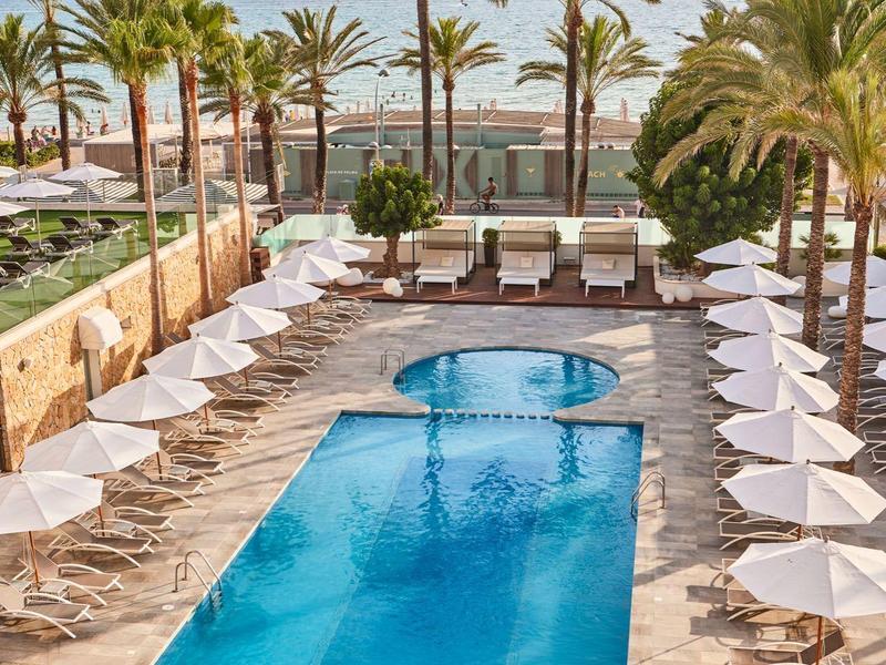 Piscina del hotel con agua azul, sombrillas blancas y palmeras en la playa.