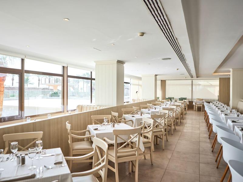 Comedor luminoso con mesas y sillas de madera junto a grandes ventanas.